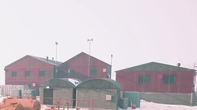 Three structures stand in the snow during winter. The buildings are surrounded by snow and have green roofs. The area looks empty and cold with a foggy sky above.
