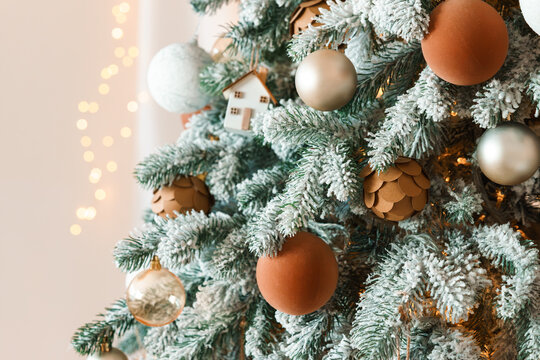 Close up of snow covered Christmas tree branch decorated with rustic brown, white and gold ornaments, miniature houses and warm lights, with soft bokeh on background. Holiday mood, festive atmosphere
