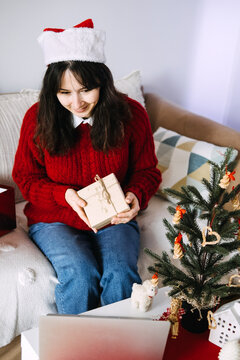 Young woman in Santa hat holds wrapped gift while smiling at laptop screen during video call at home. long distance couple christmas, virtual date december, missing partner holidays