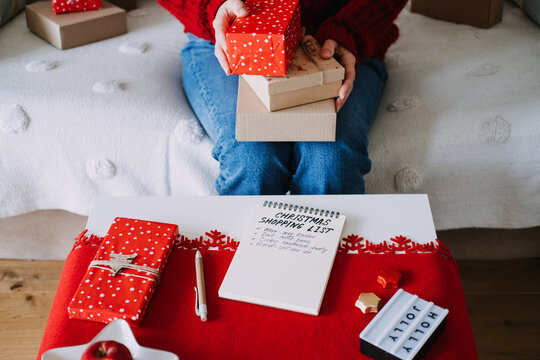 Woman holds stack of wrapped boxes above table with CHRISTMAS SHOPPING LIST notebook and HOLLY JOLLY sign. Christmas gift list planning, personalized gifts for family, mindful holiday shopping