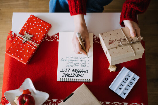 Woman writes in notebook with CHRISTMAS SHOPPING LIST on table beside wrapped gifts and HOLLY JOLLY sign. Early christmas shopping, shopping before black friday, holiday season preparation