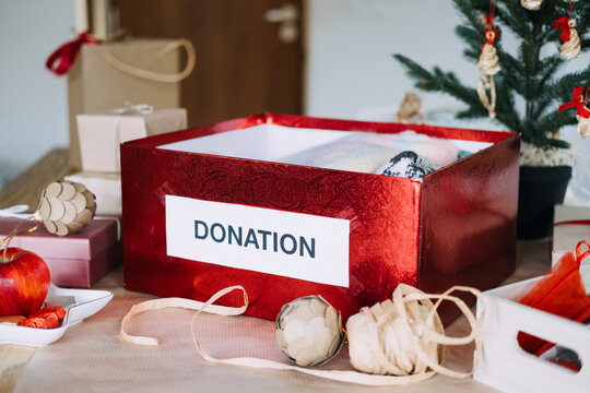 DONATION box packed with knitwear stands on kitchen table among wrapped gifts near Christmas tree. Decluttering before christmas, minimalism holiday season, conscious consumerism gift giving