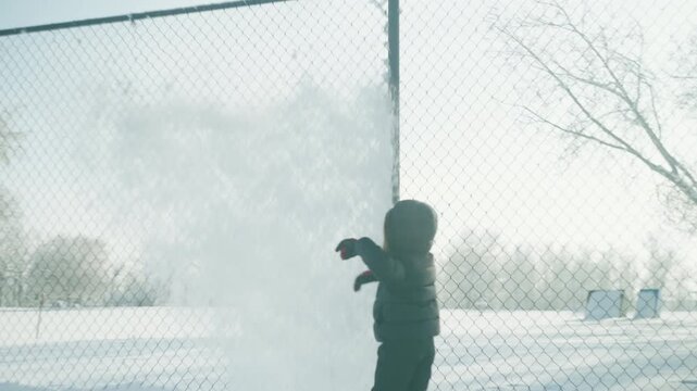Wide shot of a toddler standing by a chain-link fence in a snow-covered park with soccer nets in the distance