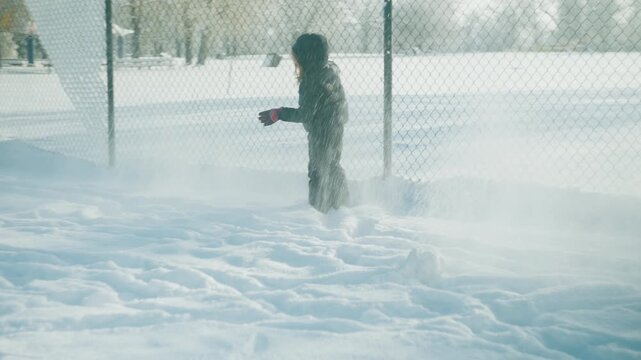 Toddler in dark snowsuit walking through deep snow near a chain-link fence on a bright winter day