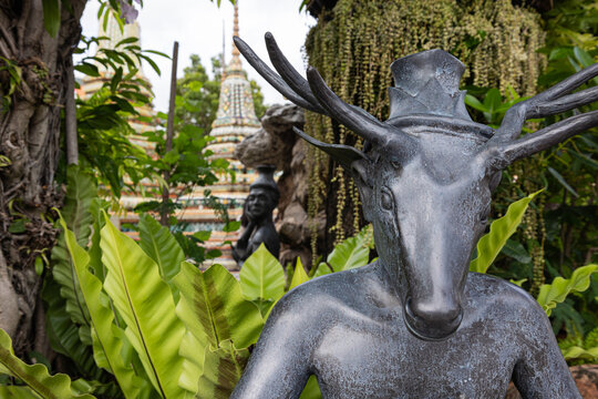 Das Bild zeigt eine dunkle Metallstatue eines anthropomorphen Wesens mit einem Hirschkopf und einem Geweih im Tempelgarten des Wat Pho in Bangkok.