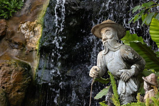 Die Abbildung zeigt eine traditionelle Steinstatue im Wat Pho in Bangkok eines &auml;lteren Mannes mit Bart und Hut, die an einer k&uuml;nstlichen Felswand mit Wasserfall  platziert ist.