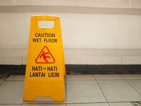 Yellow caution wet floor plastic sign on tiled floor, bilingual safety warning sign in English and Indonesian