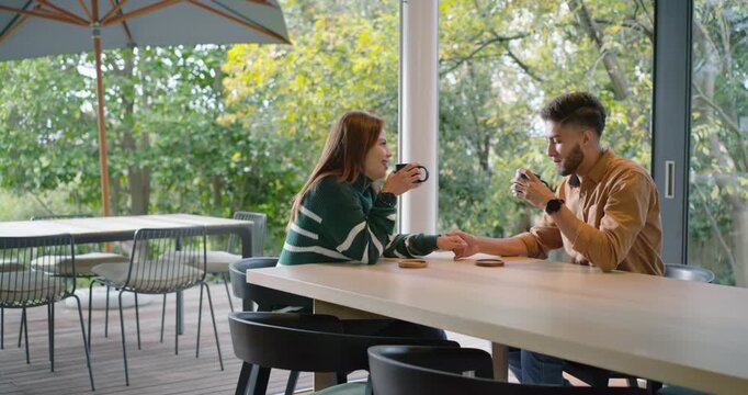 Couple at table with mugs, cam, talking, woman in green sweater reaching for man's hand, comforting