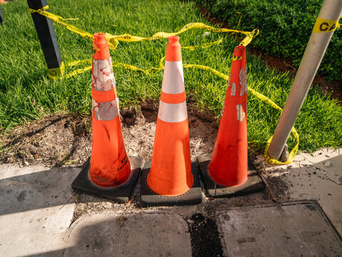 Traffic construction cones are standing on the asphalt pavement near the grass lawn boundary with the caution tape on the outdoor street.