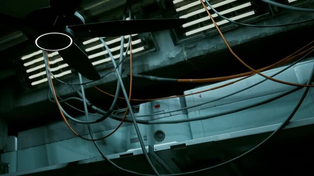 A fan spins on the ceiling in a dim room. Wires hang down from the ceilings and walls. The metal surfaces reflect light and create shadows. The space looks industrial and functional.