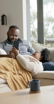 Vertical video: African American couple on sofa under throw, woman reaching for phone to read text