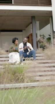 Vertical video: African American couple sitting on porch steps with mugs while man turning, talking
