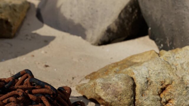 A rusty chain rests on the sand near various stones. The scene shows a beach with sunlight shining on the sand and rocks. The area looks natural with no people around.