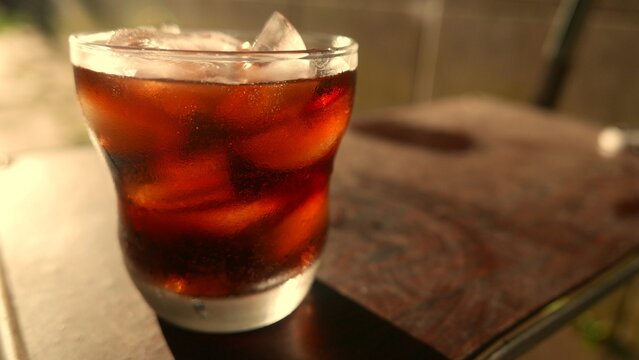 Close-up: a large soda glass under the sun. A cola soda with ice. A glass of soda on the table. POV of a soda glass. Soda with ice cubes. Iced soda. A soft drink. A fizzy drink. A pop. A refreshment. 
