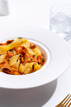 Fresh mushroom pasta dinner in bright light. Summer seasonal harvest and traditional gastronomy concept. Minimalist serving of delicious fettuccine in plate with water glass and gold fork.