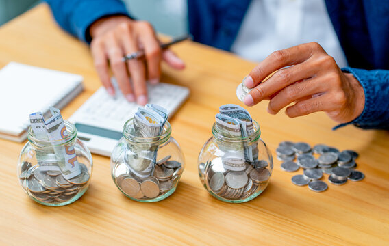 Person depositing coin into glass jar labeled 'PURPOSE' among three jars with rolled bills and coins on wooden desk