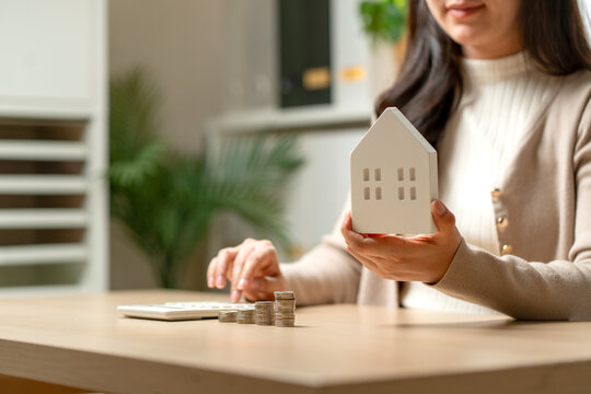 Woman holding white house model while calculating mortgage with coins and calculator on wooden desk