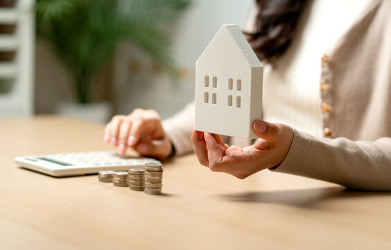 Woman holding white house model while calculating mortgage with coins and calculator on wooden desk