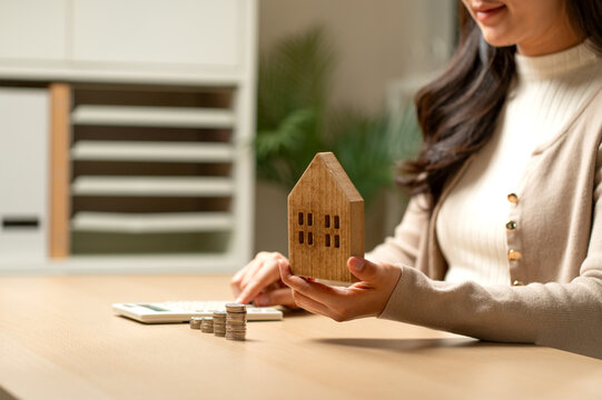 Woman holding wooden house model while calculating mortgage with coins and calculator on desk