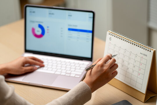 Person reviewing financial dashboard on laptop with pen poised beside standing desk calendar