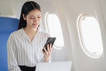 Fototapeta premium Asian businesswoman talking on smartphone while sitting in airplane cabin near window