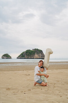 Happy Asian mother holding baby son celebrating first birthday with number 1 balloon on beach.