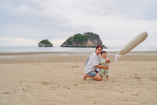 Happy Asian mother holding baby son celebrating first birthday with number 1 balloon on beach.