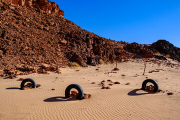Wide landscape view of discarded car tires resting on sand dunes near rocky hills © Buğra Kaan Ersoy