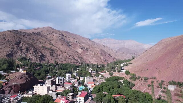 North Road village occupies a valley beside a reddish mountain wall, houses and trees under a bright highland sky. Ideal for travel, tourism, and nature stock Iran Alborz mountains valleys.