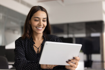 Obraz premium Smiling young 35s Arabian businesswoman holds digital tablet in modern office, enjoying online content or participate in video call, review new business platform, respond to corporate clients remotely