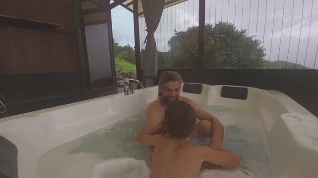FPV drone aerial reveals father and son relaxing in bubbling hot tub of geodesic glamping dome in Costa Rica surrounded by green rainforest hills and misty clouds on cloudy tropical day.