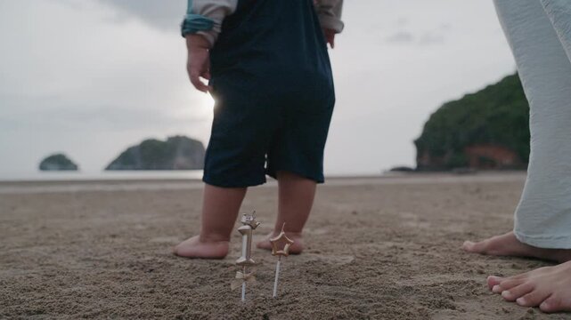 Asian mother holding hands with toddler son walking on sandy beach together.