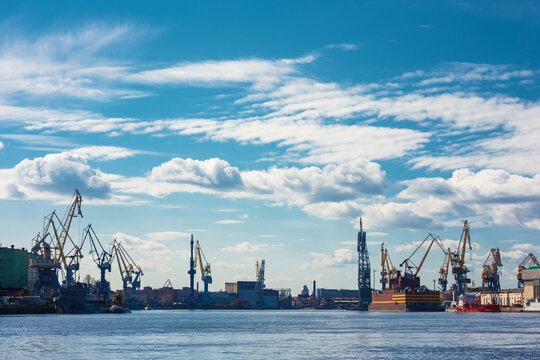 Cranes and vessels line the Admiralty Shipyards along the Neva River in Saint Petersburg beneath a blue sky with scattered clouds, showcasing heavy shipbuilding infrastructure