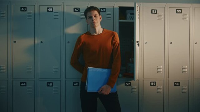 Confident High School Student in Orange Long Sleeve Shirt Stands by Metal Lockers Holding Blue Folder and Looking at Camera While Classmates Prepare For Lessons in Bright School Hallway. High School.