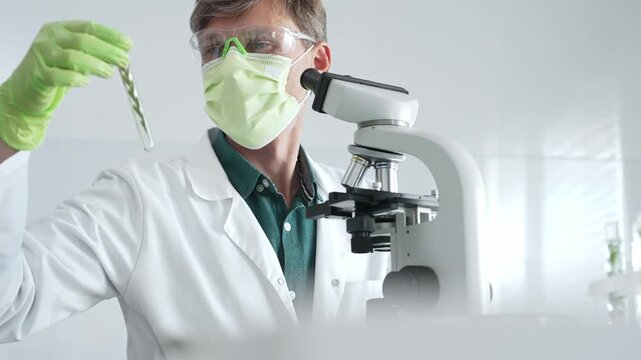 Adult male laboratory researcher wearing green protective gear examining green liquid sample through microscope during medical experiment. Medicine, healthcare and science concept