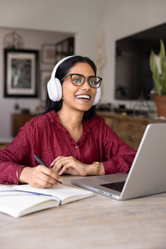 Young Indian woman wearing headphones sits at home with laptop, smiling and taking notes in notebook, engaged in online lesson or virtual meeting, enjoying communication and remote learning. E-learn