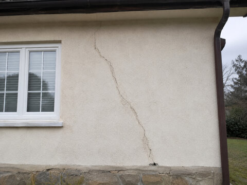 Riss in der verputzten Au&szlig;enwand eines Wohnhauses neben einem Fenster und Fallrohr, sichtbar bis zum Sockelbereich.