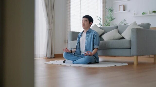 Happy young Asian man practicing yoga and meditation at home sitting on floor in living room in lotus position and relaxing with closed eyes. Mindful meditation and wellbeing concept