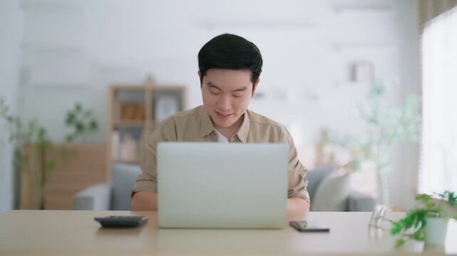 Happy young Asian man working on laptop in living room. Freelance worker male sitting at desk writing notes while watching webinar, studying online