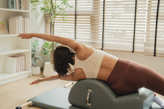 Rear view of a woman stretching sideways on a Pilates arc barrel, hands clasped behind her head, one leg extended, in a sunlit studio with wooden floors.