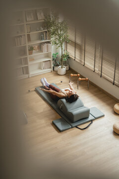 Overhead view of a woman lying on a Pilates arc barrel with arms raised toward the ceiling, in a warm sunlit studio with a fireplace, bookshelf, and wooden fitness props nearby.