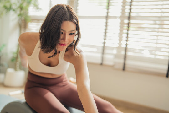Woman performing a seated forward stretch on a Pilates arc barrel, reaching toward her extended foot, in a sunlit studio with wooden floors and green plants.