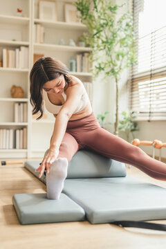 Woman performing a seated forward stretch on a Pilates arc barrel, reaching toward her extended foot, in a sunlit studio with wooden floors and green plants.