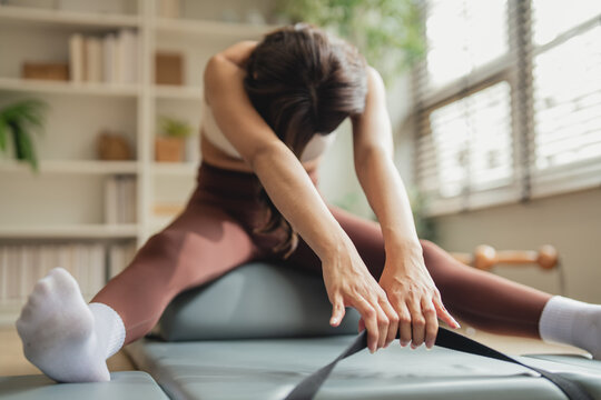 Close-up of a woman performing a deep forward split stretch on a Pilates barrel, using a resistance strap to extend her reach, soft natural light in a home studio.