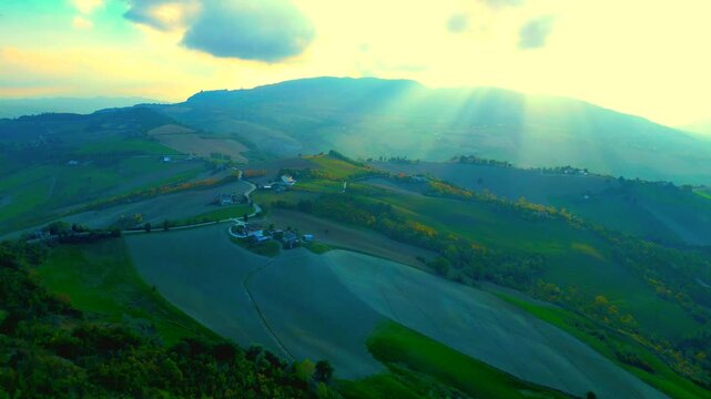 Glorious aerial pullback shot from near Santa Vittoria in Matenano, the stunning light of the sun piercing through the clouds and the Sibillini Mountains, imbuing the sky with golden immortal quality
