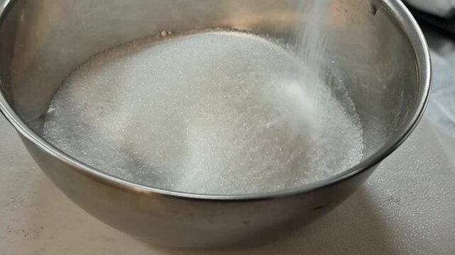 Pouring white granulated sugar into a stainless steel bowl.
