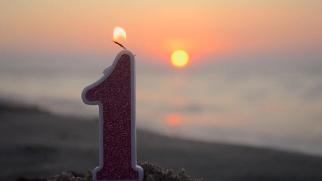 Numeral one from paraffin wax with pink glitters burns on sandy beach of sea shore at dawn sunset close-up. Paraffin candle burning near blue sea. Concept holiday Birthday New Year party celebrating