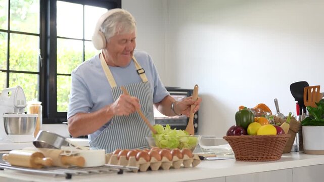 Happy senior man listening to music with headphones cooking in kitchen, elderly man wearing headphones and cooking salad with wife at home