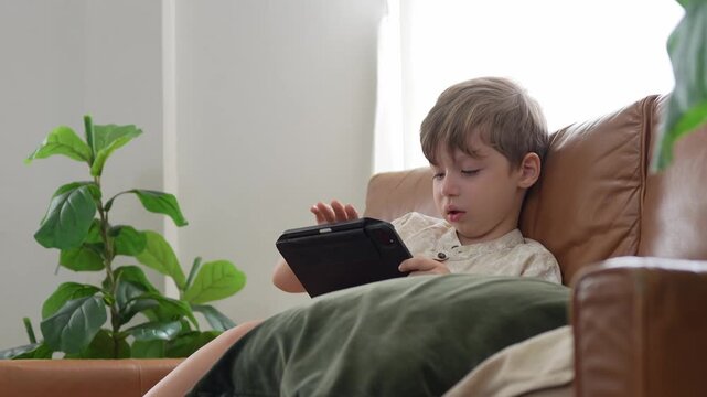 Kid boy using digital tablet sitting on sofa at home, little boy playing game on tablet computer in living room, child looking at tablet screen and relaxing on couch indoors