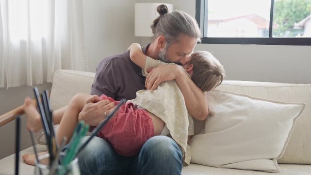 Father hugging and kissing his little son on sofa at home, dad embracing and playing with son in living room indoors, father and son sharing tender moment and cuddling together on couch